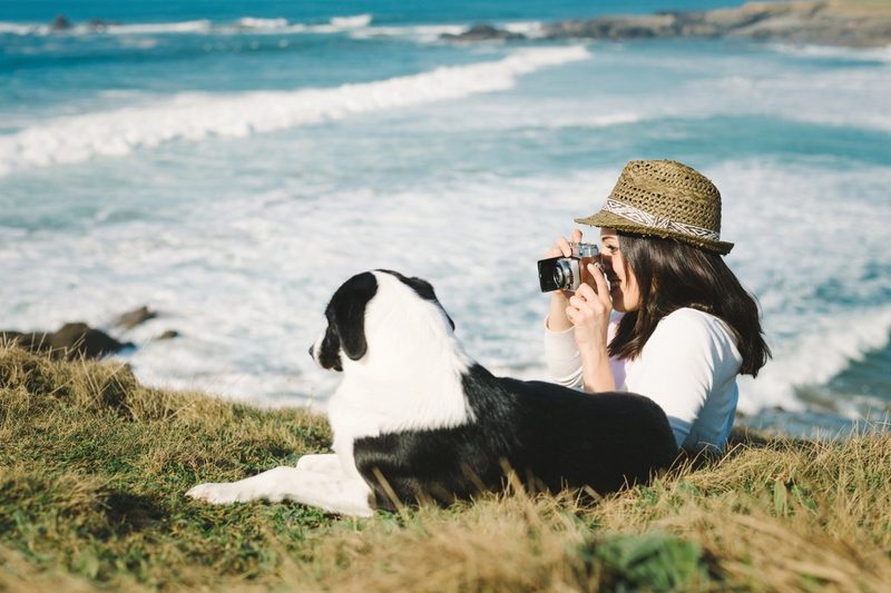 Voyageuse prenant une photo au bord de la mer avec son chien allongé dans l’herbe, lors d’un séjour en pleine nature.