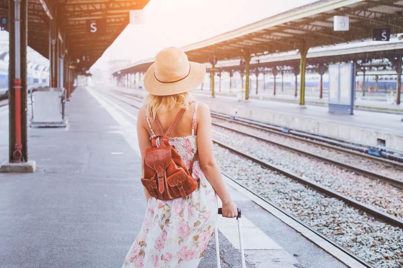 Une femme portant une robe à motifs floraux et un chapeau de soleil marche le long d'un quai de gare avec une valise. Le soleil matinal éclaire la scène, donnant une ambiance de début de voyage.