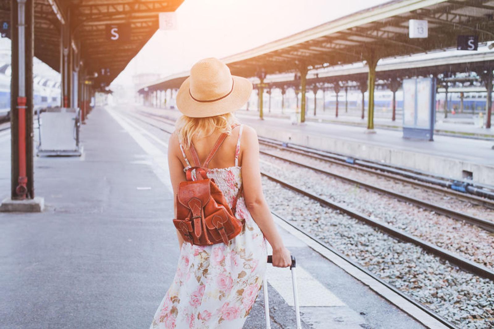 Une femme portant une robe à motifs floraux et un chapeau de soleil marche le long d'un quai de gare avec une valise. Le soleil matinal éclaire la scène, donnant une ambiance de début de voyage.