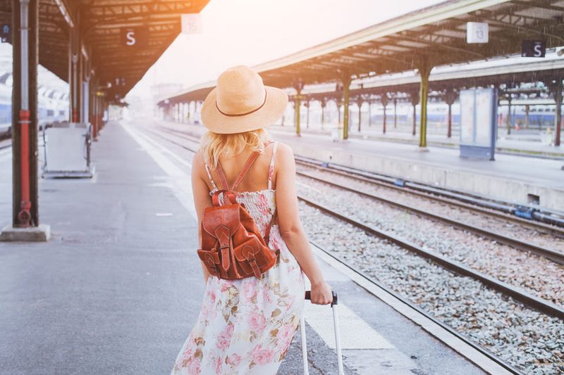 Une femme portant une robe à motifs floraux et un chapeau de soleil marche le long d'un quai de gare avec une valise. Le soleil matinal éclaire la scène, donnant une ambiance de début de voyage.