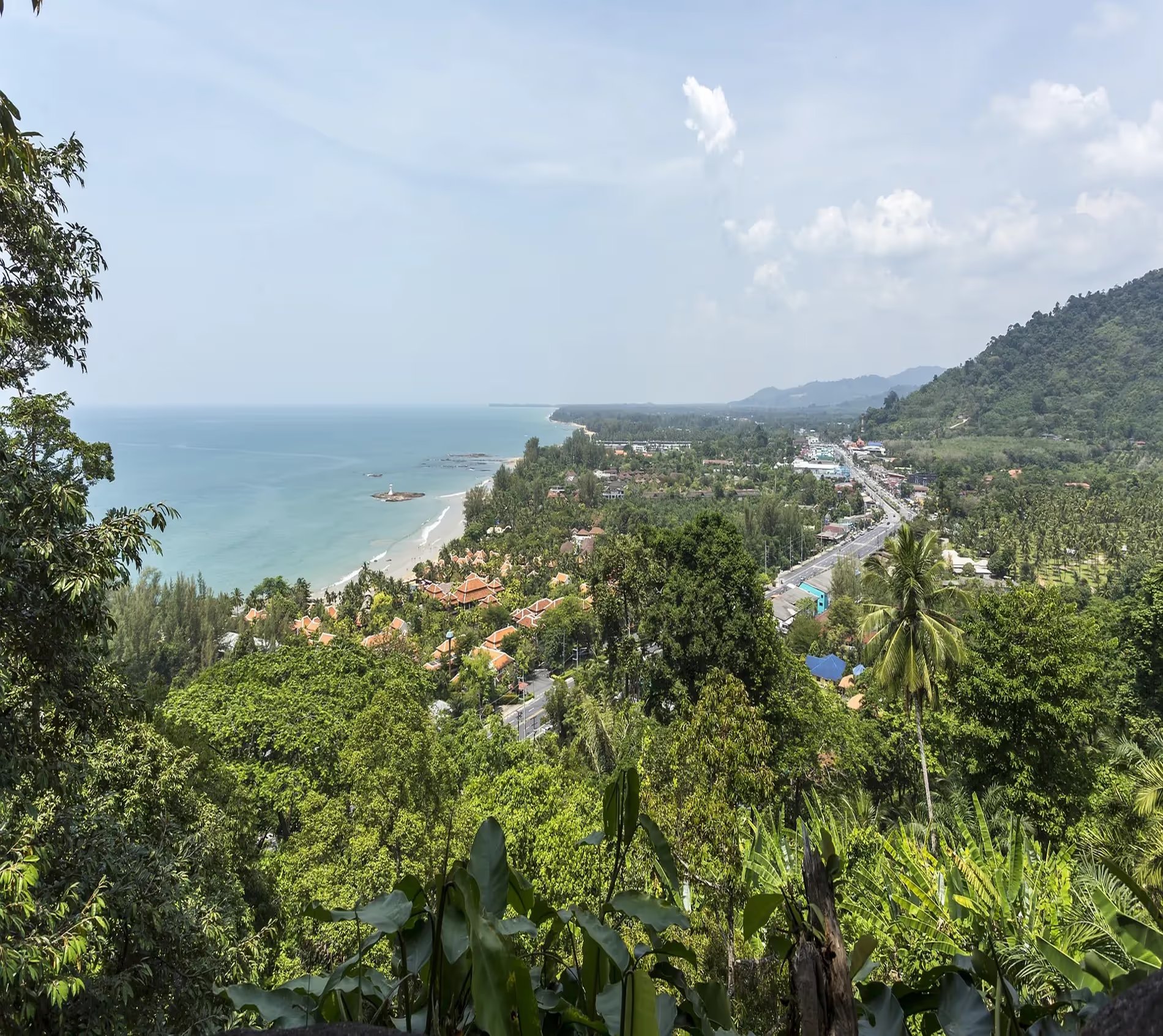 Vue panoramique de Khao Lak, montrant la côte avec la mer turquoise, la plage, les bâtiments avec des toits rouges et la végétation luxuriante sous un ciel légèrement nuageux.