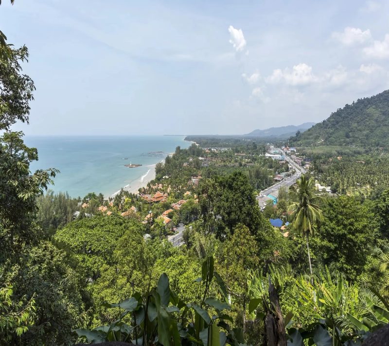 Vue panoramique de Khao Lak, montrant la côte avec la mer turquoise, la plage, les bâtiments avec des toits rouges et la végétation luxuriante sous un ciel légèrement nuageux.