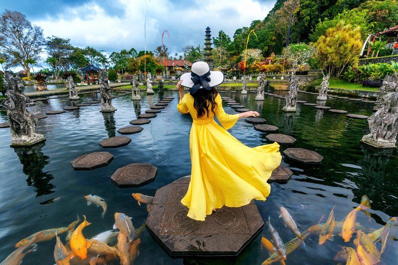 Femme en robe jaune marchant dans le jardin de Tirta Gangga sur les pierres, entourée d’étangs et de statues.