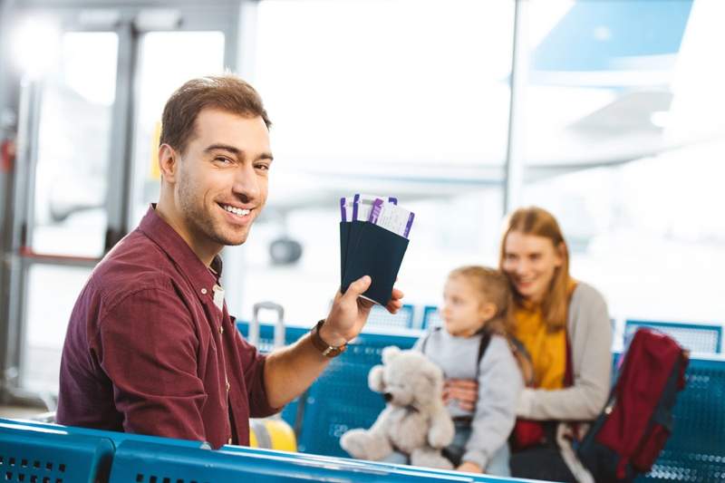 Famille avec enfant assise dans un aéroport, un père souriant tenant des billets d’avion avant un voyage en avion.