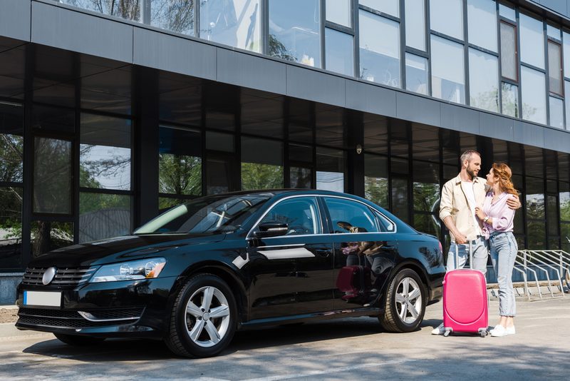 Couple avec une valise récupérant leur voiture de location noire devant une agence de location