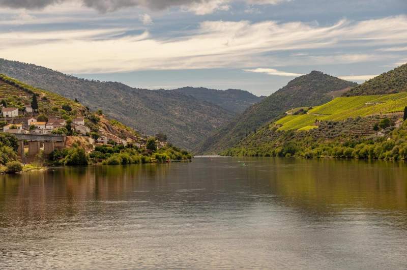 Paysage de la vallée du Douro au Portugal, avec des collines verdoyantes, des vignobles et un village au bord du fleuve sous un ciel partiellement nuageux.