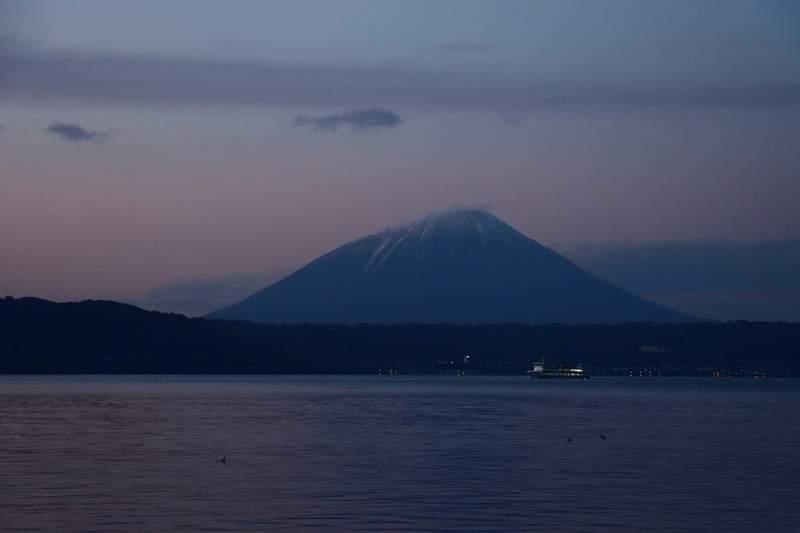 Mont volcanique enneigé au coucher du soleil reflété sur un lac calme, avec un ferry éclairé au loin.