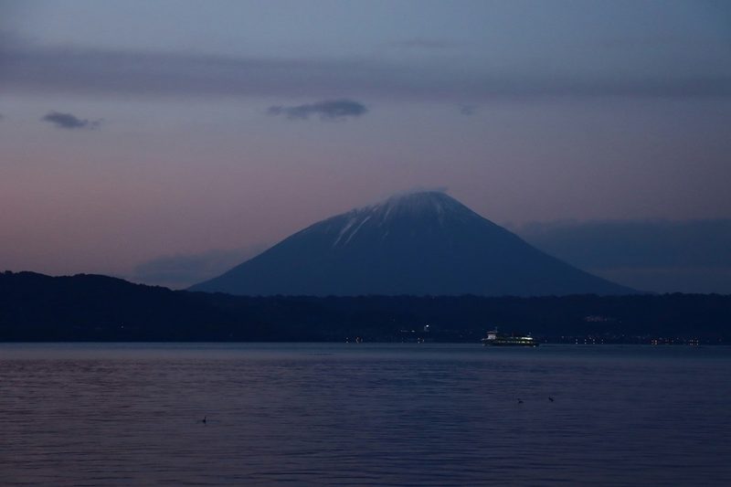 Mont volcanique enneigé au coucher du soleil reflété sur un lac calme, avec un ferry éclairé au loin.