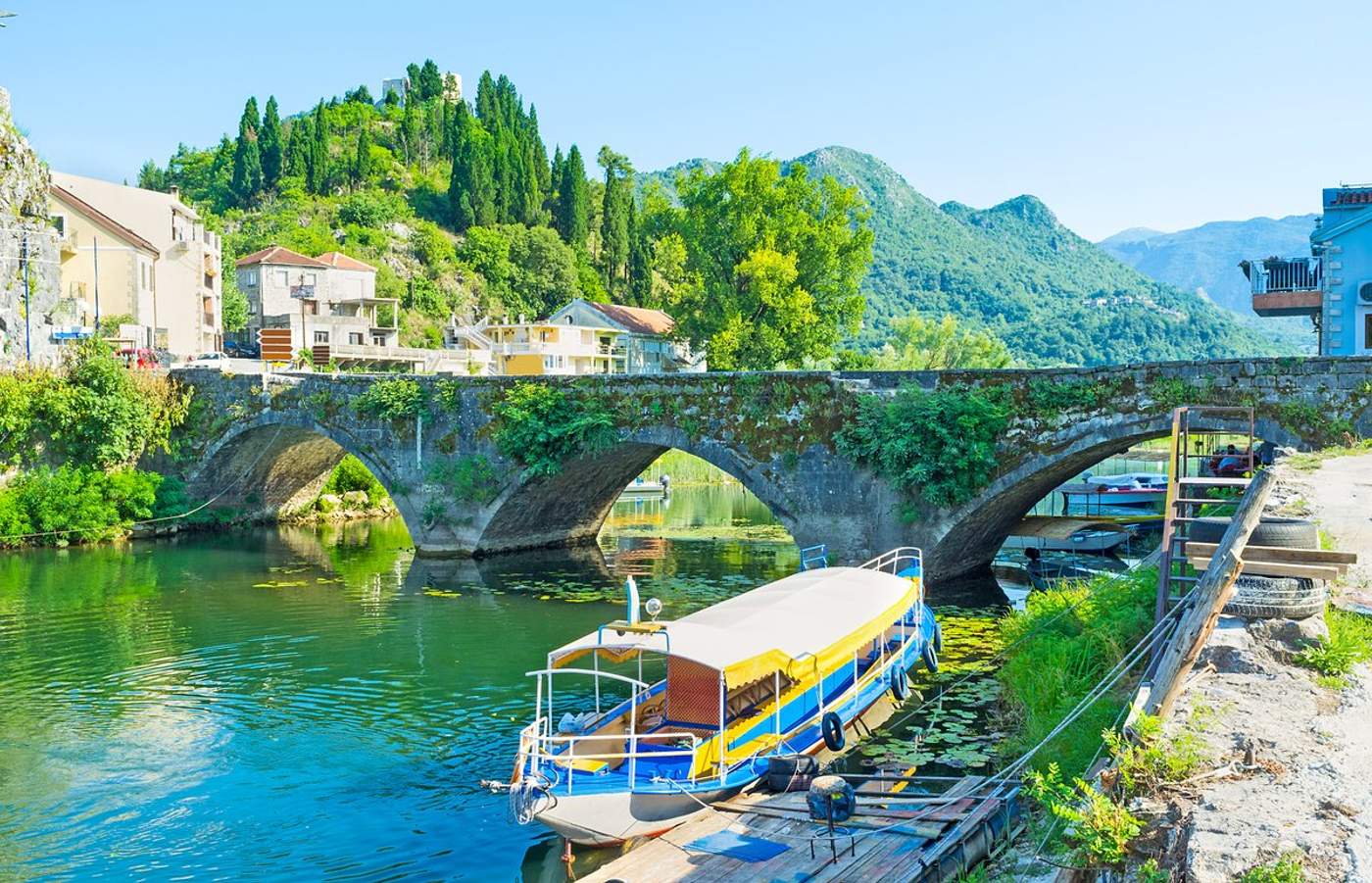 Bateau d'excursion bleu et jaune amarré sous le pont de pierre de Virpazar au Monténégro, avec le village et les montagnes verdoyantes en arrière-plan