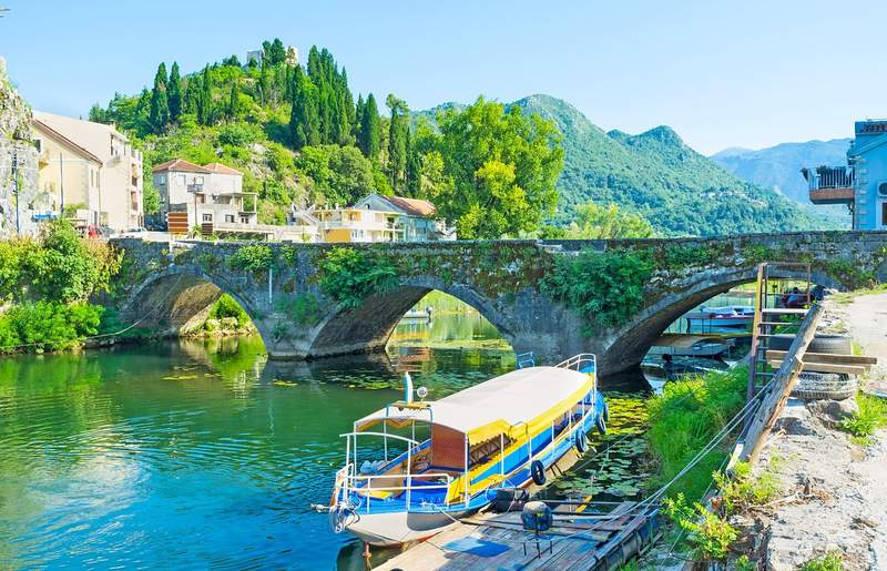 Bateau d'excursion bleu et jaune amarré sous le pont de pierre de Virpazar au Monténégro, avec le village et les montagnes verdoyantes en arrière-plan