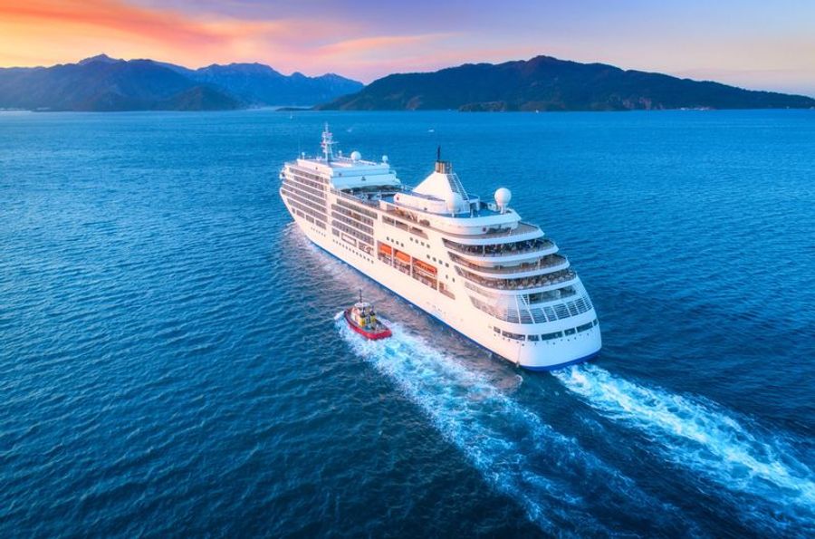 Vue aérienne d’un paquebot de croisière de luxe naviguant en mer au coucher du soleil, avec des montagnes en arrière-plan.