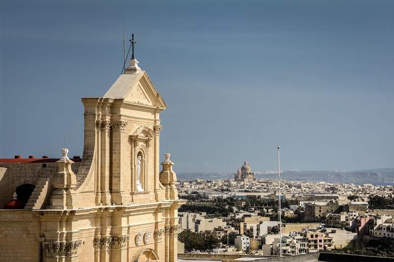 Façade de la Citadelle de Victoria à Gozo avec vue lointaine sur la basilique Ta’ Pinu et les paysages urbains maltais.