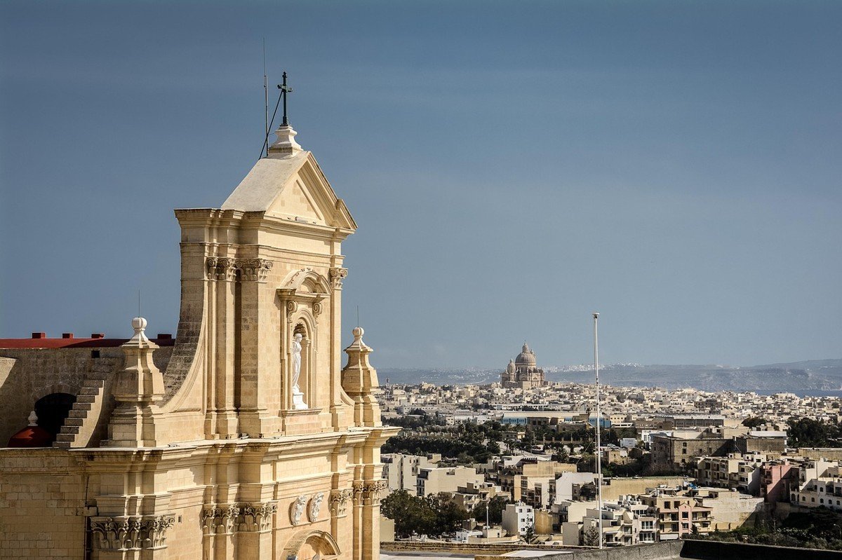 Façade de la Citadelle de Victoria à Gozo avec vue lointaine sur la basilique Ta’ Pinu et les paysages urbains maltais.