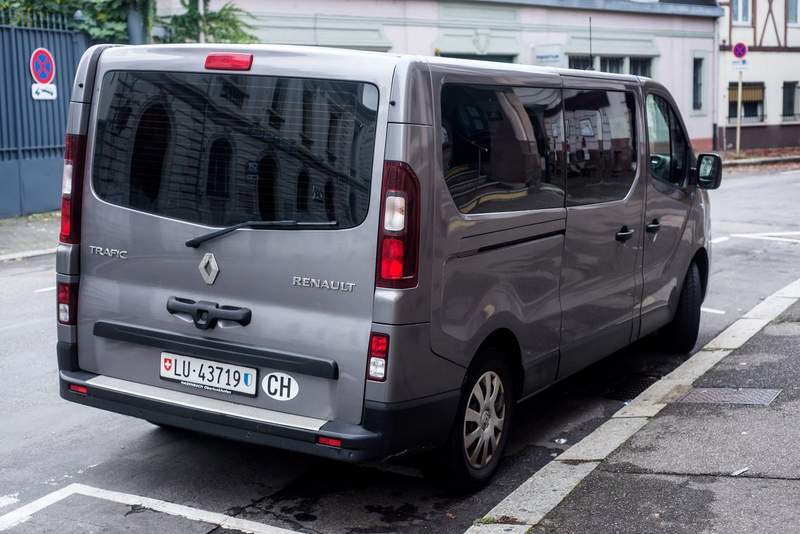 Vue arrière d’un van Renault Trafic gris métallisé, immatriculé en Suisse, stationné dans une rue urbaine pavée.
