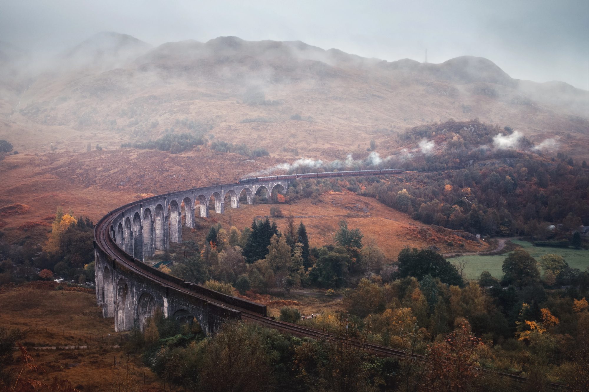 Un train à vapeur et ses voitures rouges franchissent le viaduc courbe de Glenfinnan, dans une vallée aux couleurs d’automne sous la brume