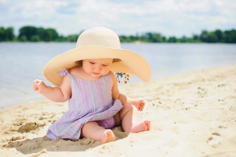Bébé assis sur le sable avec un grand chapeau, au bord d’un lac par une journée ensoleillée.