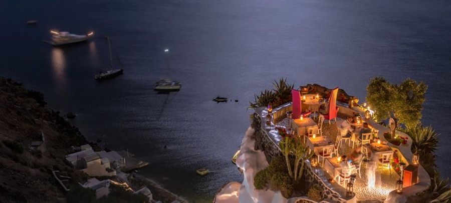 Terrasse illuminée à Mykonos avec vue sur la mer éclairée par la lune et des bateaux au loin.
