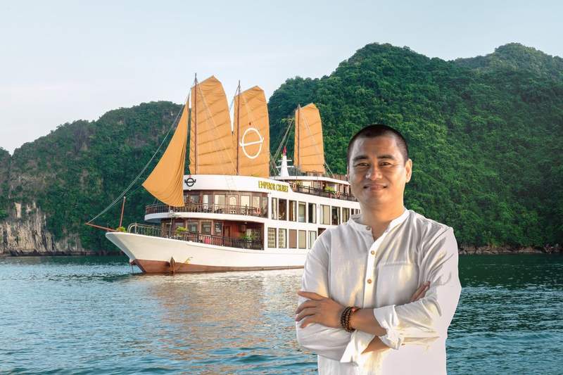 Homme souriant en chemise blanche, bras croisés, devant un jonque traditionnelle Emperor Cruises voiles ocre déployées dans la baie d'Ha Long