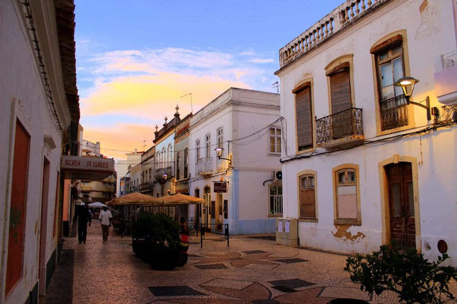Une rue piétonne bordée de bâtiments traditionnels portugais avec des façades colorées et des balcons en fer forgé, illuminée par une lumière de fin de journée.