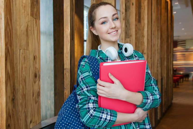 Une étudiante souriante tenant des livres et portant un sac à dos dans un couloir moderne d'université ou d'une école.
