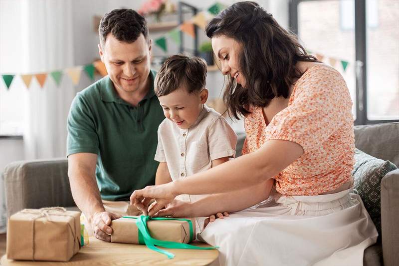 Un enfant ouvre un cadeau en famille, accompagné de ses parents souriants, dans un salon décoré — moment chaleureux avant un départ en voyage.