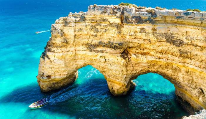 Vue aérienne d'une formation rocheuse emblématique de l'Algarve, au sud du Portugal, avec un bateau traversant les eaux turquoise en dessous d'une arche naturelle en pierre.