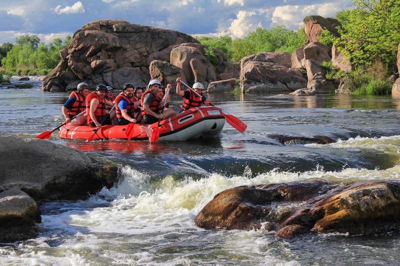 Groupe en gilets de sauvetage rouges faisant du rafting sur une rivière bordée de rochers, sous un ciel partiellement nuageux, pagaies rouges en main.