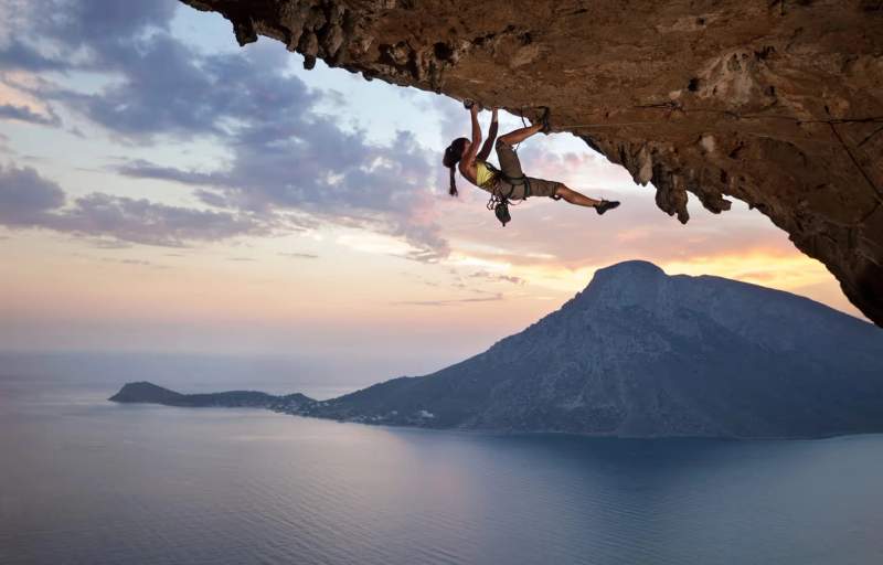 Escalade d'une falaise au coucher du soleil avec une vue panoramique sur la mer et les montagnes en arrière-plan.