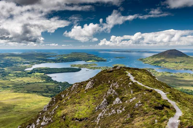 Vue panoramique depuis les hauteurs du Connemara, montrant des sentiers sinueux, des lacs scintillants et des îles au loin sous un ciel bleu parsemé de nuages.