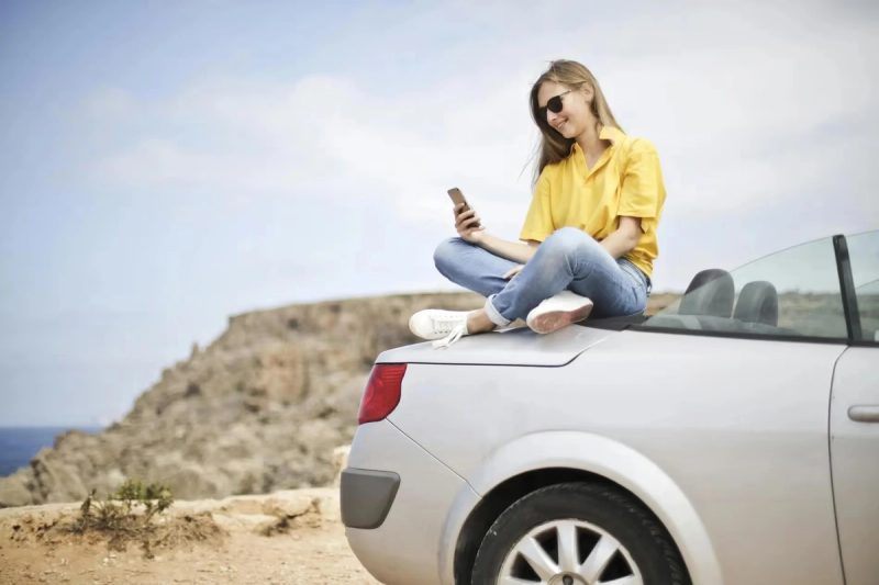 Femme en chemisier jaune et jean bleu prenant un selfie assise sur une voiture