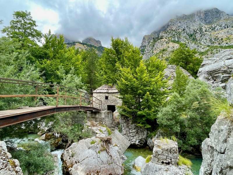 Pont en bois traversant une rivière entourée de végétation luxuriante et de montagnes à Theth, Albanie.