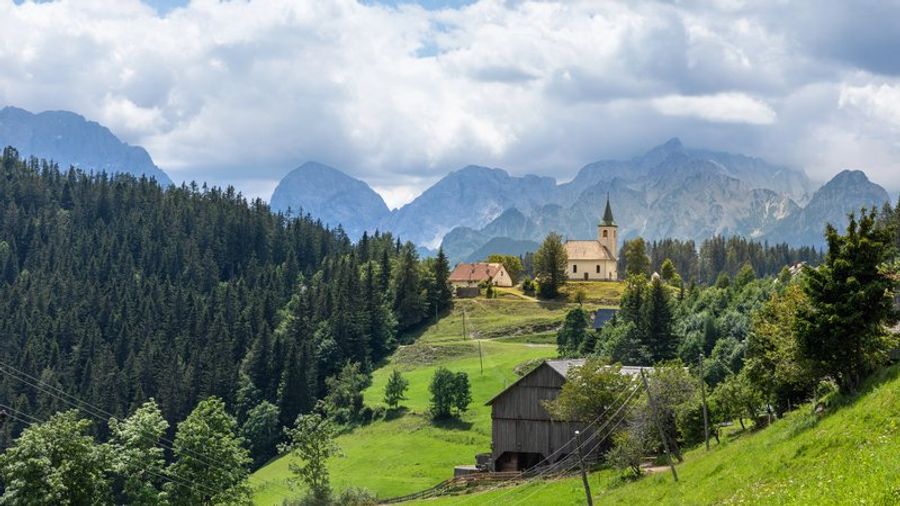 Petite église blanche sur une colline boisée en Slovénie, entourée de prairies vertes et dominée par les crêtes des Alpes Juliennes sous un ciel nuageux