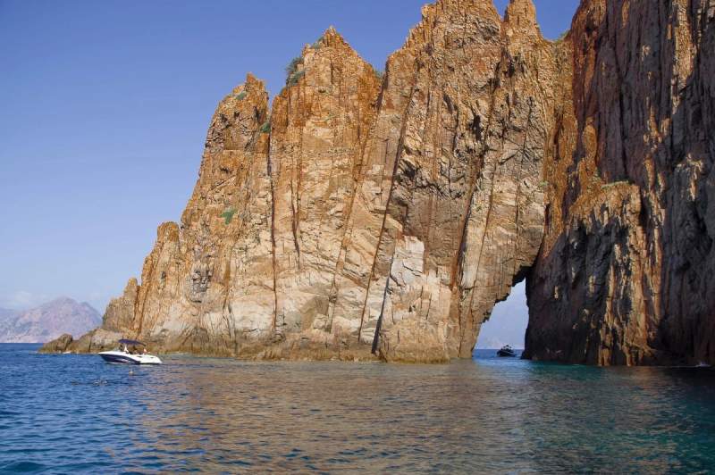 Vue spectaculaire des falaises imposantes des calanques de Corse, avec des formations rocheuses rouges plongeant dans la mer Méditerranée, et des bateaux naviguant à proximité.