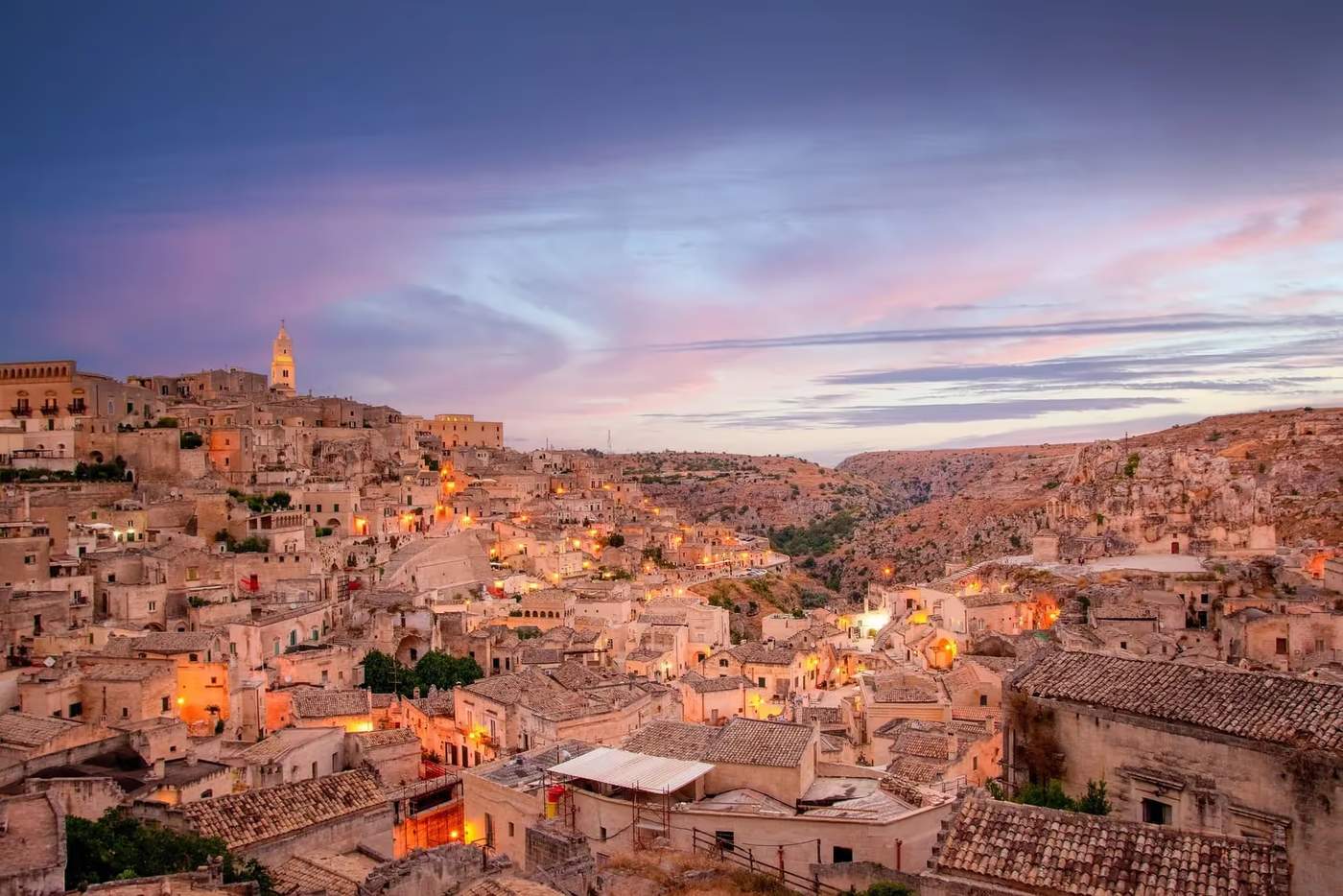 Vue panoramique de Matera avec ses bâtiments historiques en pierre, la cathédrale de Matera surplombant la ville et les habitations troglodytiques typiques, sous un ciel partiellement nuageux.