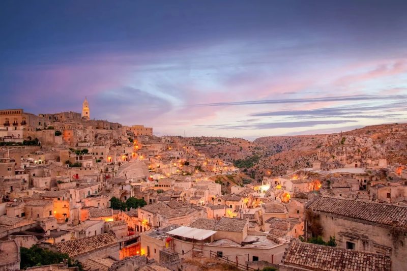 Vue panoramique de Matera avec ses bâtiments historiques en pierre, la cathédrale de Matera surplombant la ville et les habitations troglodytiques typiques, sous un ciel partiellement nuageux.