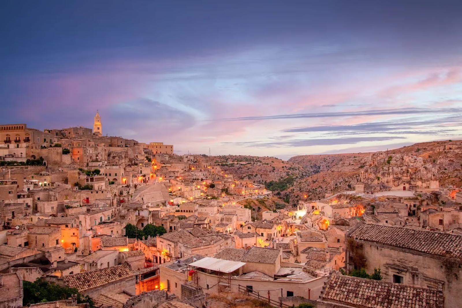 Vue panoramique de Matera avec ses bâtiments historiques en pierre, la cathédrale de Matera surplombant la ville et les habitations troglodytiques typiques, sous un ciel partiellement nuageux.