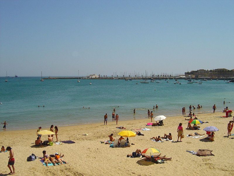 Plage de Conceição à Cascais, Portugal, avec des baigneurs profitant de la journée ensoleillée et des bateaux amarrés au loin.