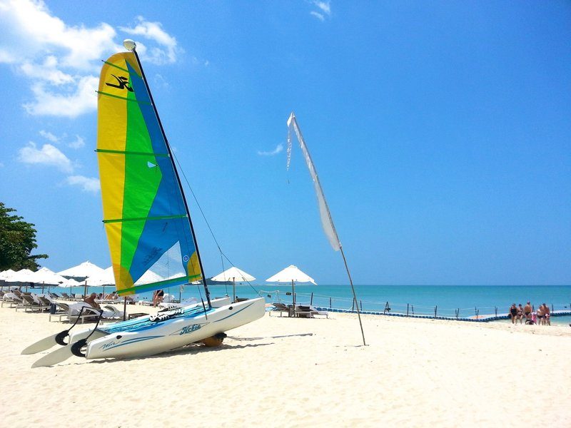 Plage de sable blanc avec un catamaran coloré et des parasols, ciel bleu dégagé.