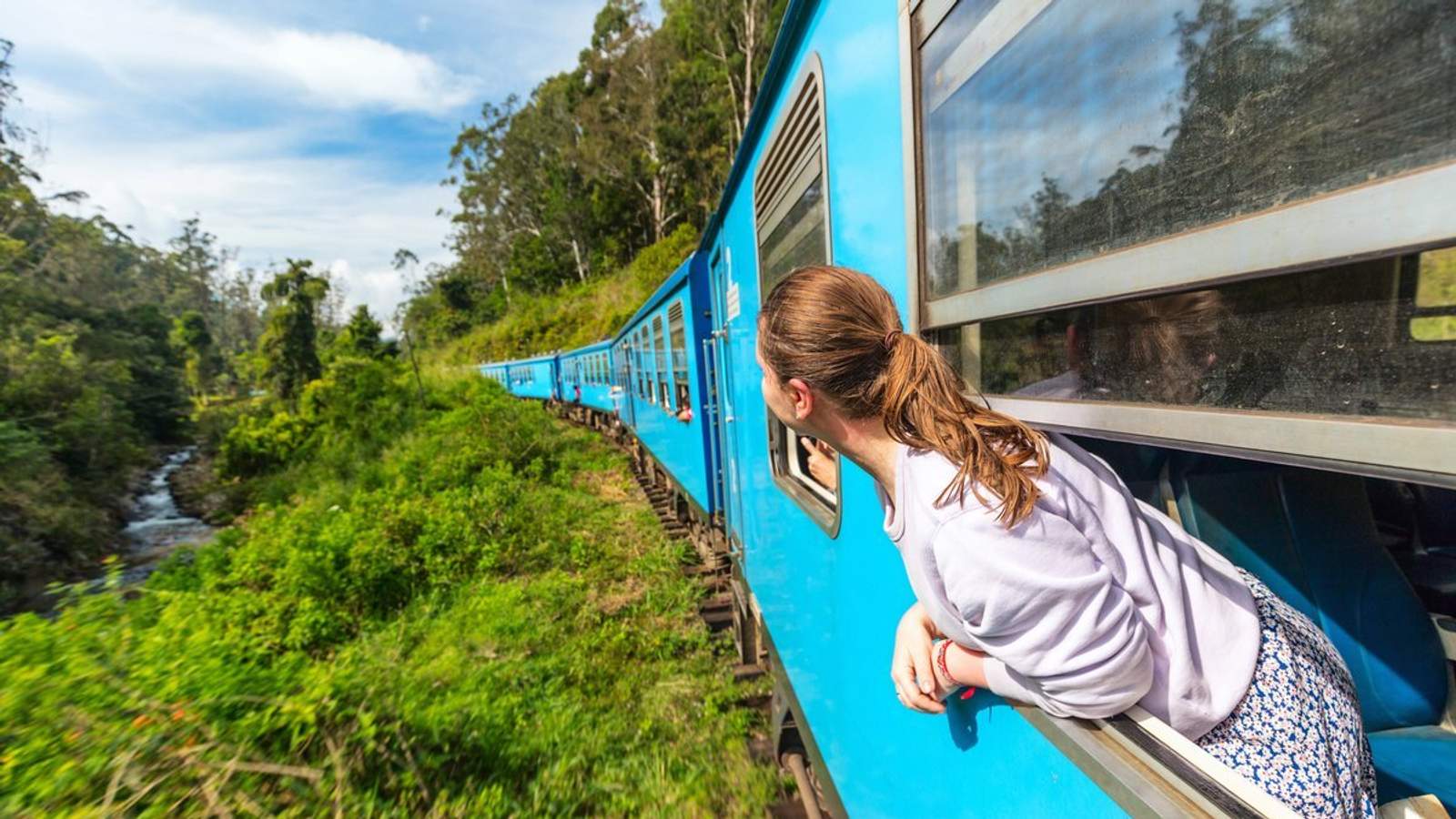 Voyageuse regardant le paysage depuis un train bleu traversant une région verdoyante, symbole du slow travel immersif.