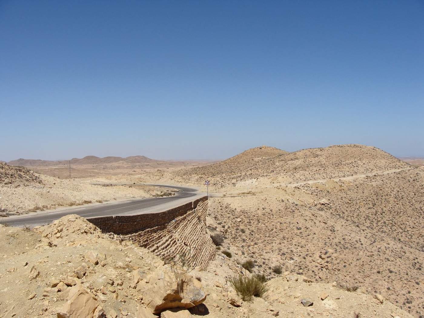 Route sinueuse traversant le désert aride du sud tunisien, entourée de collines rocailleuses ocre sous un ciel bleu dégagé