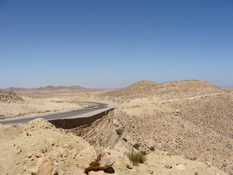 Route sinueuse traversant le désert aride du sud tunisien, entourée de collines rocailleuses ocre sous un ciel bleu dégagé