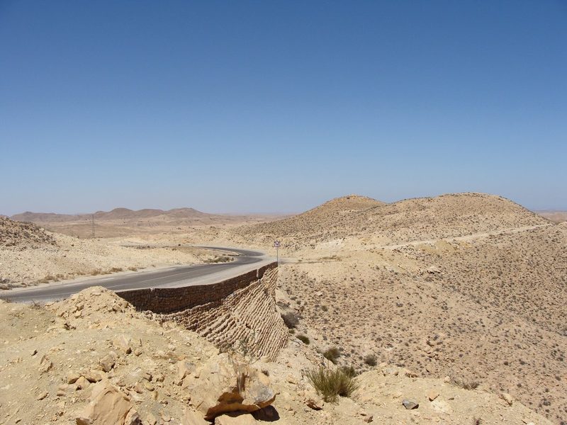 Route sinueuse traversant le désert aride du sud tunisien, entourée de collines rocailleuses ocre sous un ciel bleu dégagé