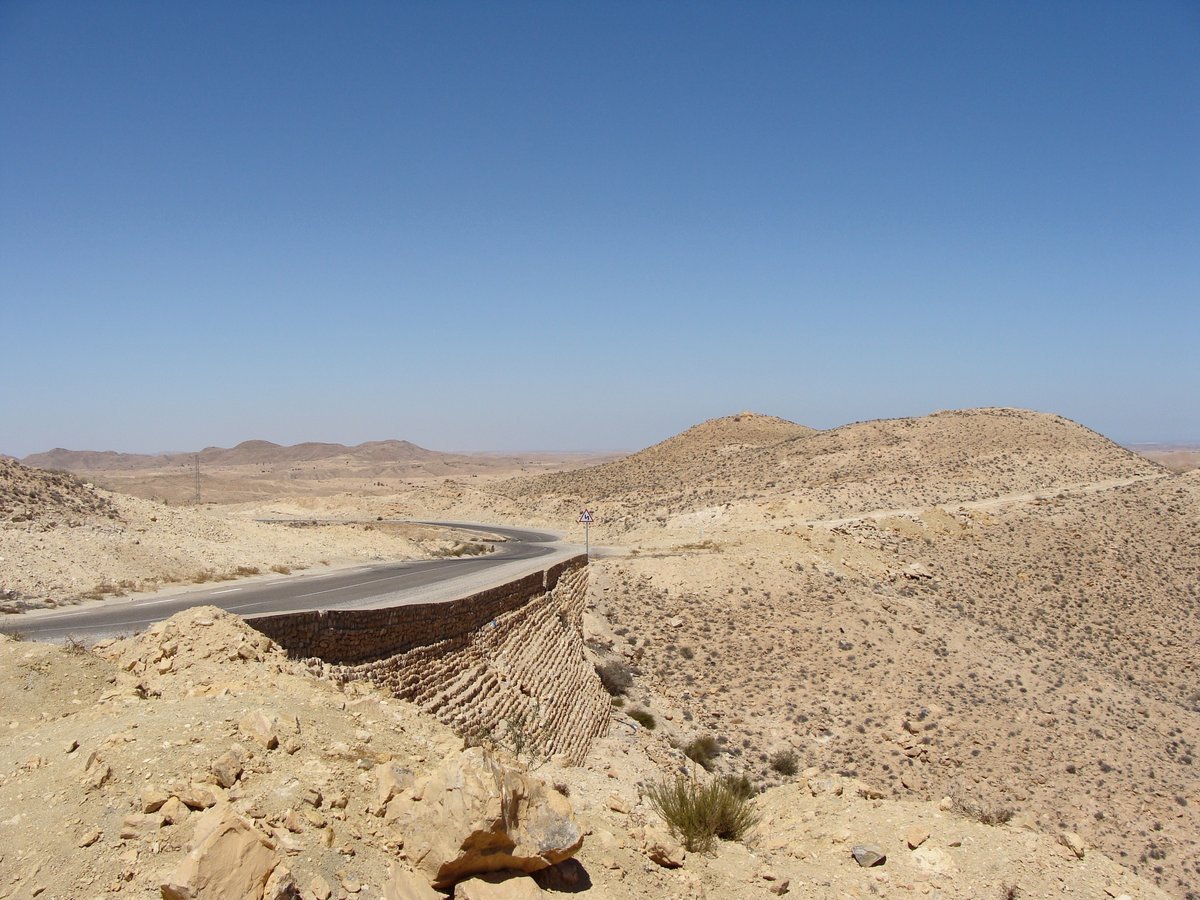 Route sinueuse traversant le désert aride du sud tunisien, entourée de collines rocailleuses ocre sous un ciel bleu dégagé