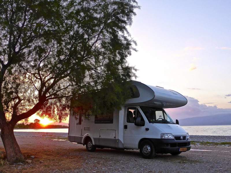 Un camping-car stationné au bord d'un lac au coucher du soleil, avec un arbre en premier plan.