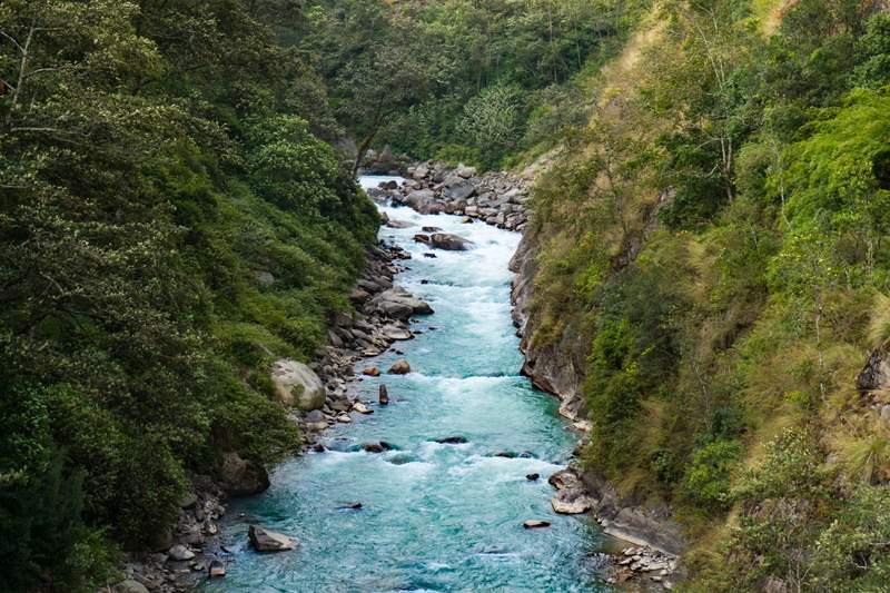 Rivière turquoise traversant une vallée verdoyante au Népal, entourée de collines boisées et de rochers.