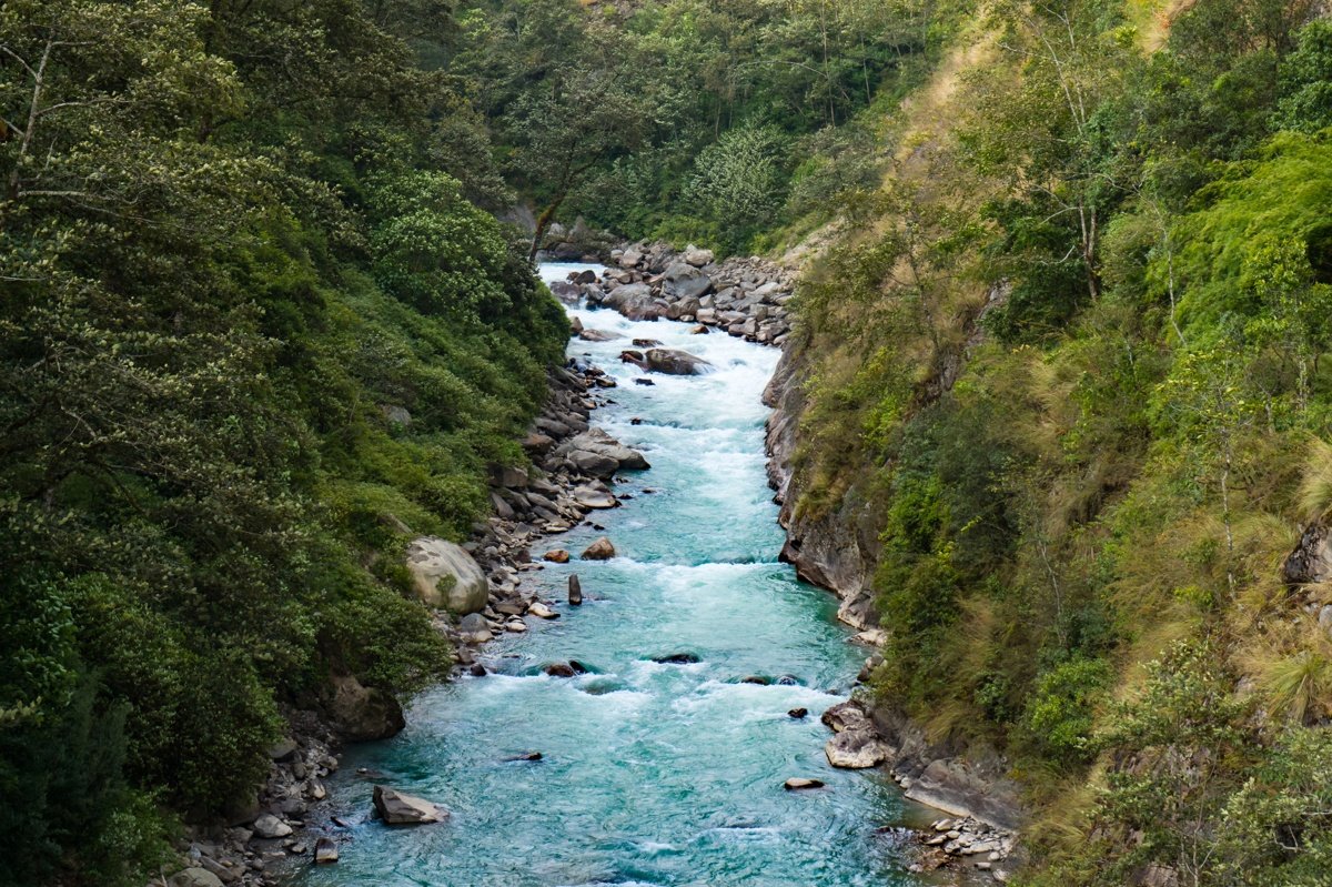Rivière turquoise traversant une vallée verdoyante au Népal, entourée de collines boisées et de rochers.