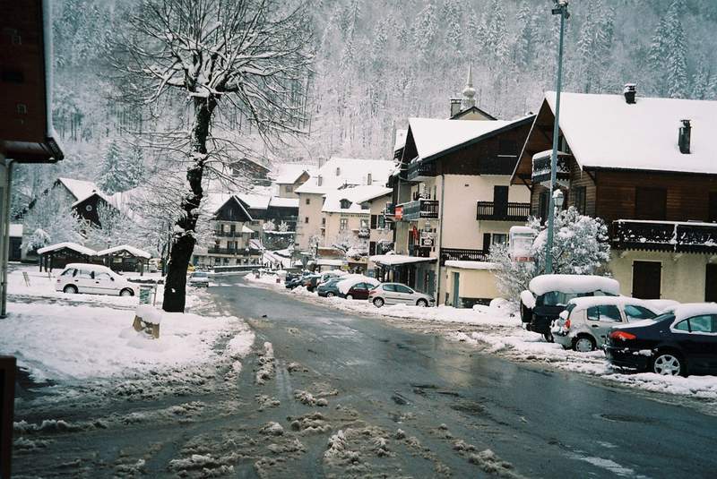 Village de montagne enneigé avec des maisons en bois, des voitures recouvertes de neige et une route partiellement dégagée en hiver.