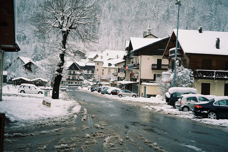 Village de montagne enneigé avec des maisons en bois, des voitures recouvertes de neige et une route partiellement dégagée en hiver.