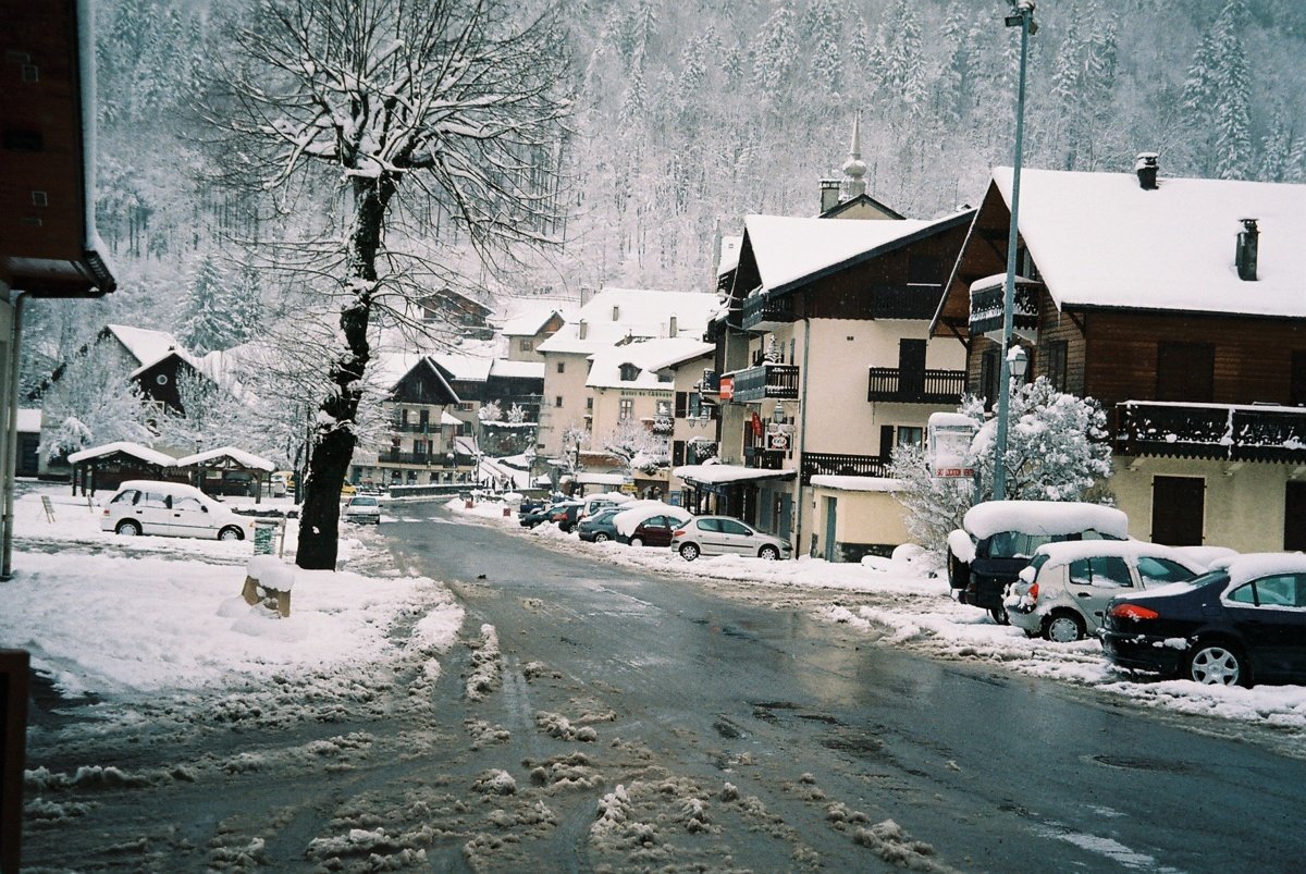Village de montagne enneigé avec des maisons en bois, des voitures recouvertes de neige et une route partiellement dégagée en hiver.