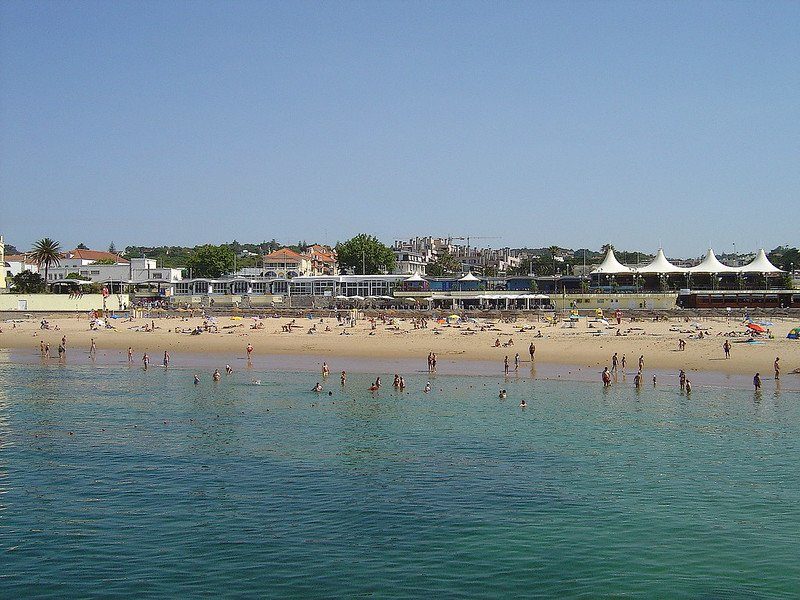 Vue de la plage de Tamariz à Cascais, Portugal, avec des baigneurs et des installations de plage en arrière-plan.